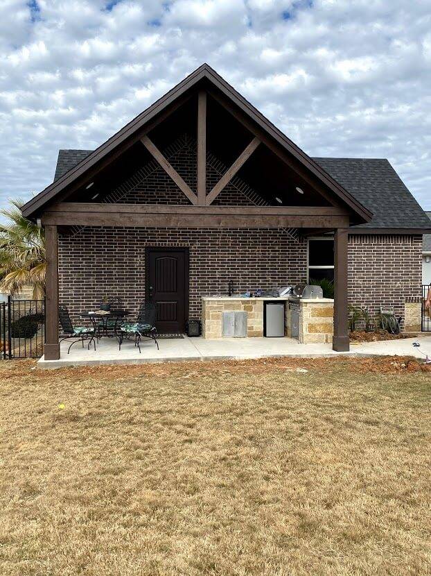 Brick home with a covered patio under a timber-framed gable, a stone outdoor kitchen and a bistro table