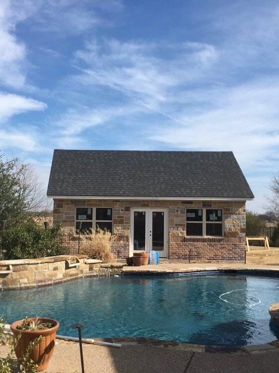 Stone and brick building with French doors and a gray shingle roof beside a swimming pool